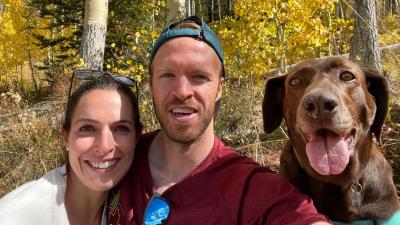 Nova the dog with mouth open in a smile with tongue out next to two people outside in the woods