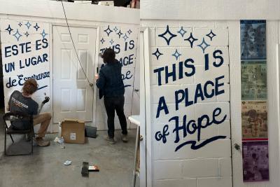Two people painting words on a wall that says, "This is a place of hope. Este es un lugar de esperanza."