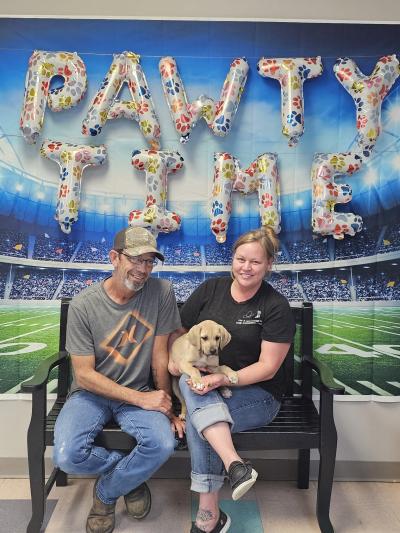 Two people sitting with a puppy under balloons forming the words Pawty Time and a football field backdrop