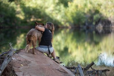 Person and dog on a rock with the person's arm hugging the dog and water and trees in the background
