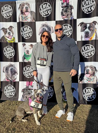 Jess and her husband with Pearl the dog in front of a One Love backdrop