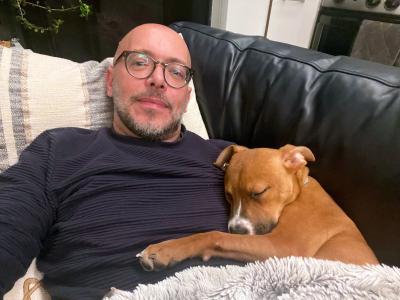 Brown dog snuggled and sleeping next to volunteer David Sprague while lying on a couch
