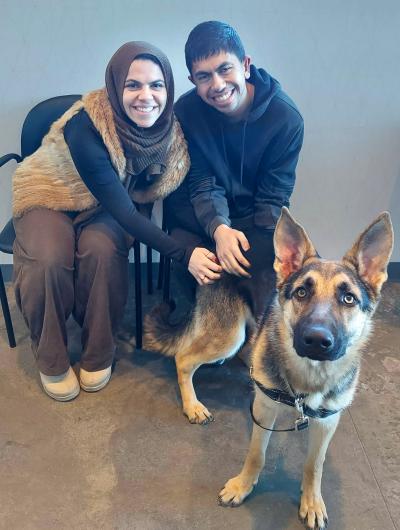 A smiling couple sitting in chairs beside the shepherd dog they just adopted