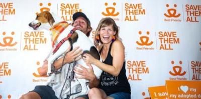 Man and woman holding tan and white hound dog in front of save them all background