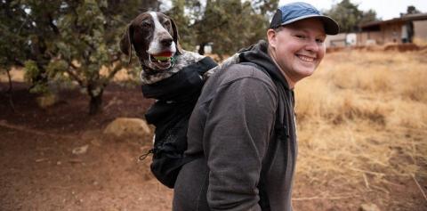 Woman in dark gray sweatshirt carrying pointer dog in black backpack