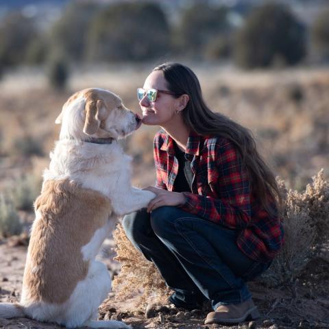 Woman in plaid shirt kneeling down outside with brown and white dog that's sitting on its hindlegs