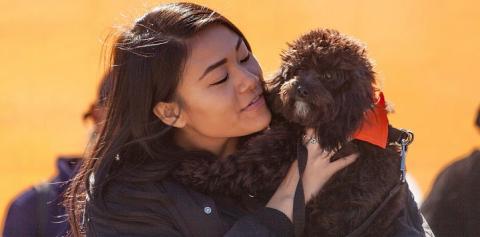 Woman with long dark hair wearing black coat holding black dog wearing orange bandana