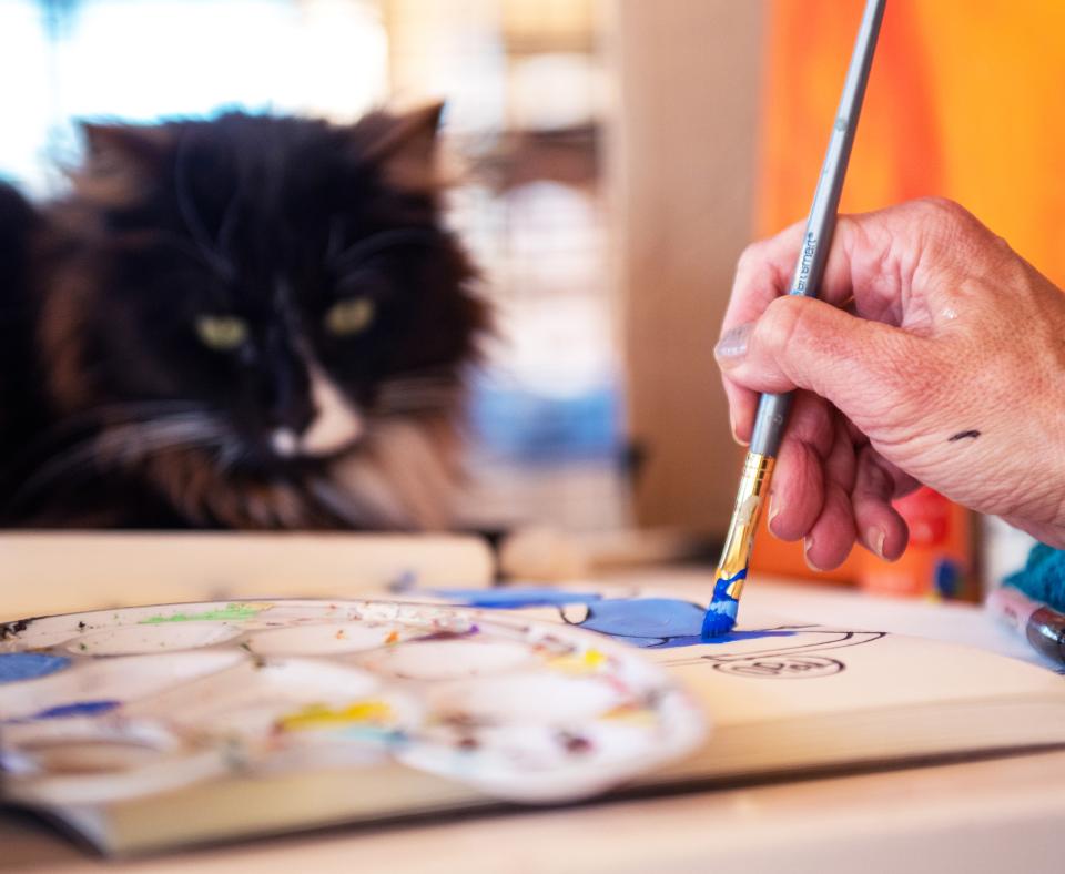 Person painting on a table while a cat looks on with interest
