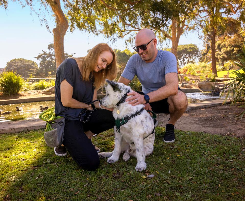 Two smiling people outside in a park kneeling down next to a dog