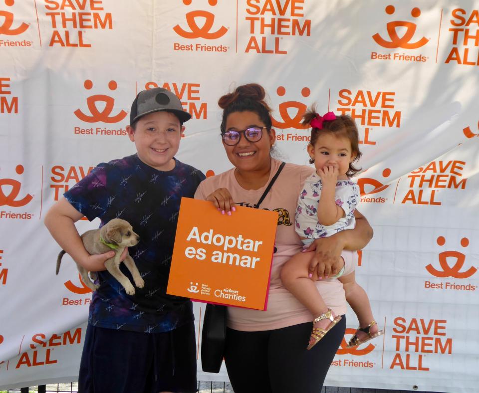 Family adopting a puppy on front of a Best Friends backdrop holding a sign that says, Adoptar es amar