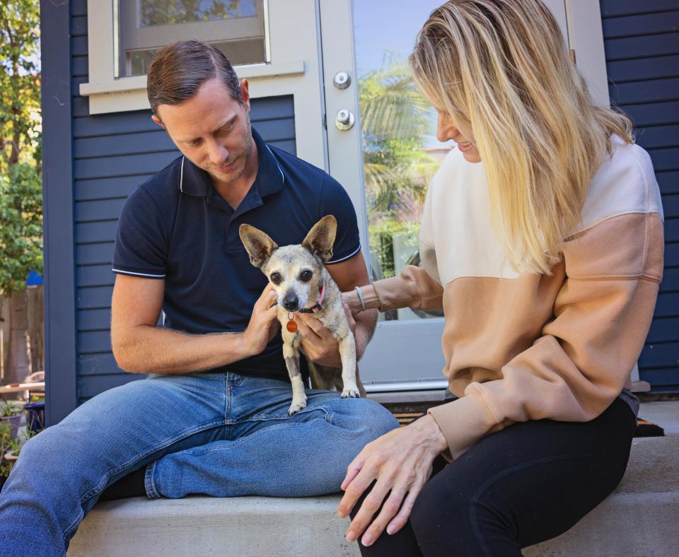 Two people sitting outside with a small dog on stairs leading into a hous
