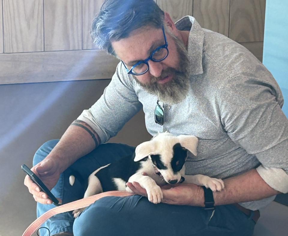 A man in glasses and a beard sits on the floor holding a black and white puppy