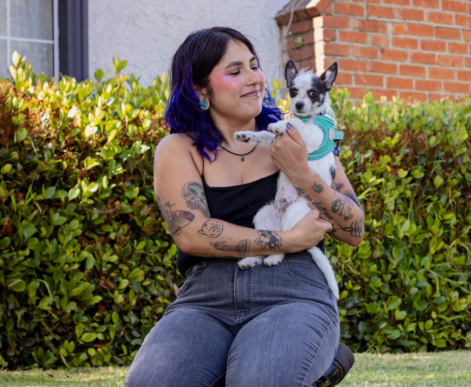 Smiling person holding a small dog while sitting in a yard next to a house