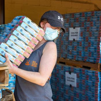 Masked person carrying a stack of cases of kitten food with pallets of food behind her