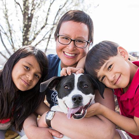 Woman and two kids hugging a smiling black and white dog whose tongue is out