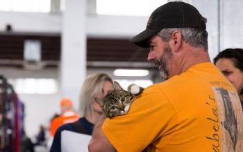 Man in black hat and yellow shirt cradling tabby cat looking at camera