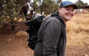 Woman in dark gray sweatshirt carrying pointer dog in black backpack