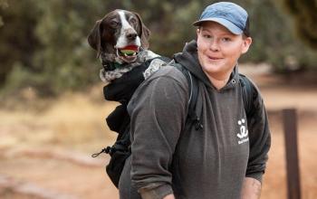 Staff member wearing hat and carrying a dog in a backpack