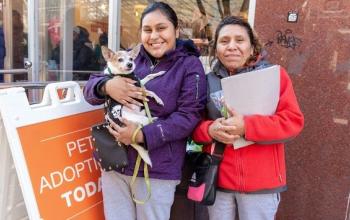 Woman in purple jacket holding small tan dog next to woman in white jacket holding a folder