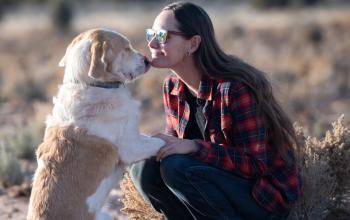 Woman in plaid shirt kneeling down outside with brown and white dog that's sitting on its hindlegs