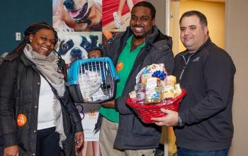Woman standing next to man holding white cat in blue carrier next to a man holding a red gift basket