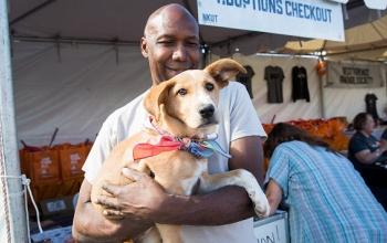 Smiling man in gray shirt holding tan dog wearing orange bandana in front of adoption booth