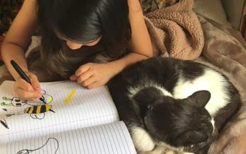 Black and white cat curled up next to little girl with dark hair writing