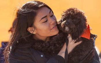 Woman with long dark hair wearing black coat holding black dog wearing orange bandana