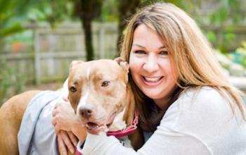 Tan pit bull standing next to sitting woman in gray top