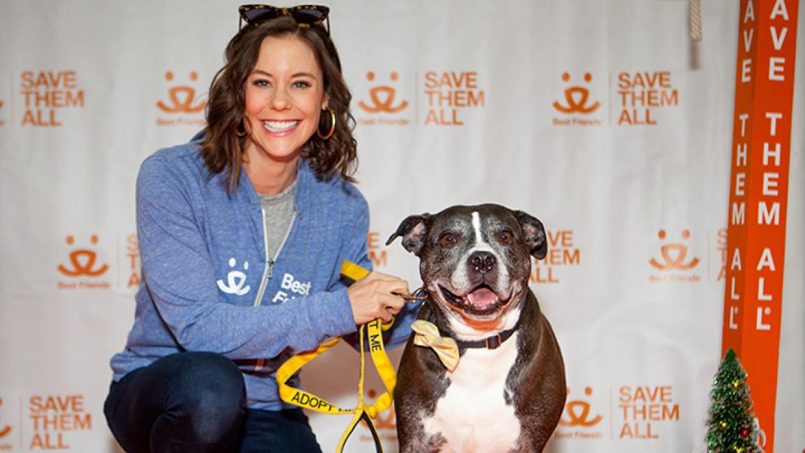Ashley Williams in front of a Best Friends backdrop, posing with a black and white pit-bull-type dog