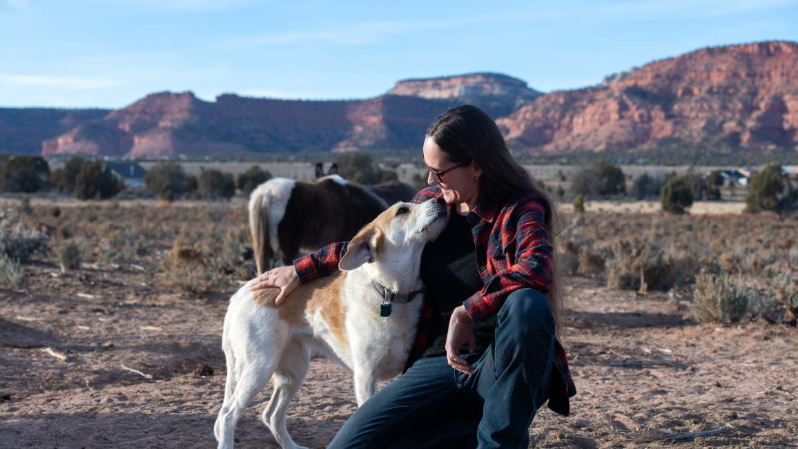 Emery holding Greenie the dog affectionately at the ranch with horses and rock formations in the background