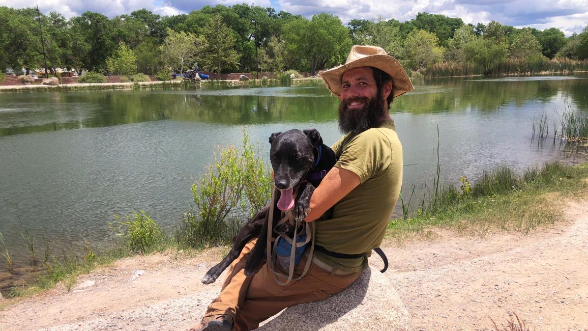 Ken wearing a cowboy hat and holding Minnie the dog in his lap, sitting on a boulder in front of a river