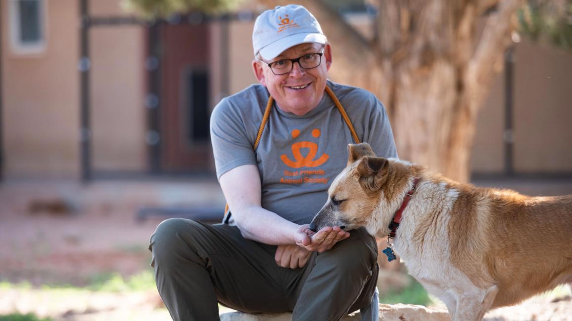 Volunteer wearing a Best Friends T-shirt sitting down and hand-feeding a treat to a dog