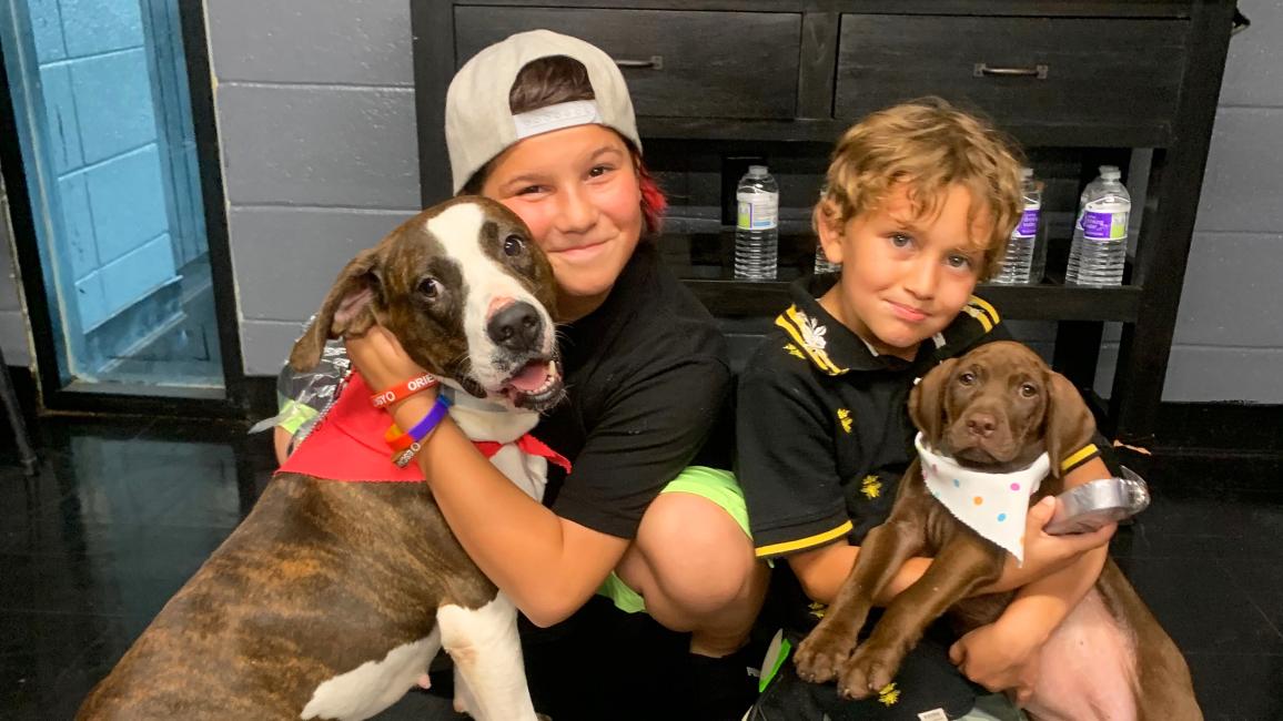 Two children associated with Grant Parish snuggling with a dog and puppy, both dogs wearing bandannas