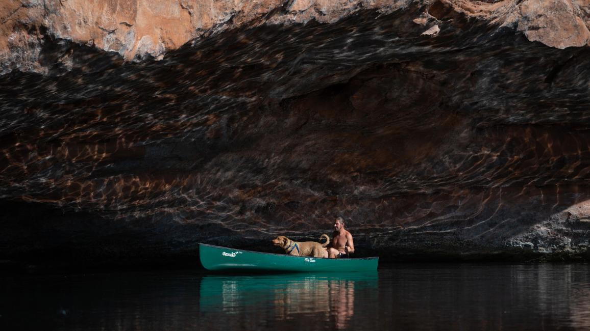 A person and dog in a green canoe on the water in front of a rock overcropping