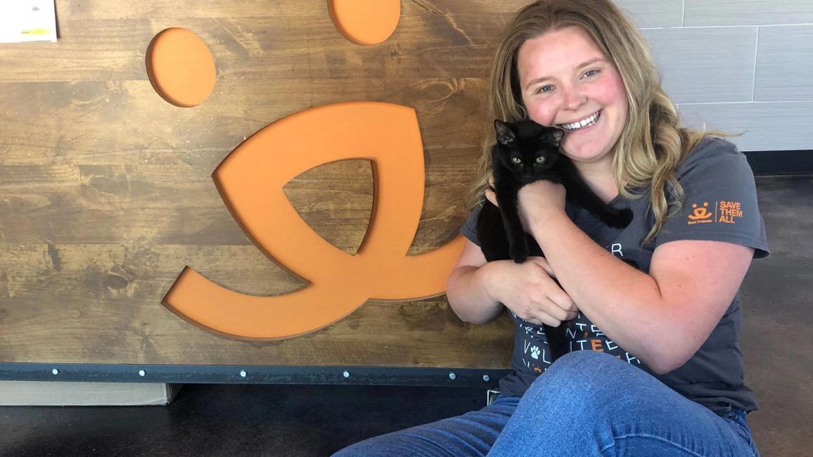 Woman holding a black cat sitting on the ground next to a Best Friends logo on the wall