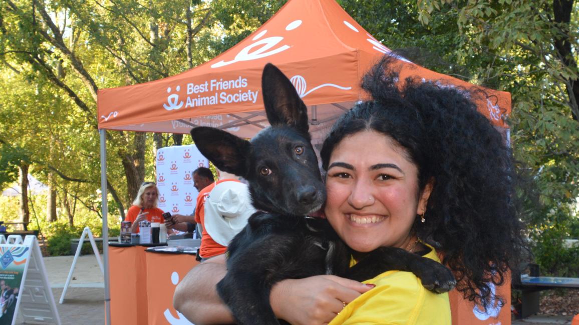 Smiling person holding the black puppy she adopted in front of a Best Friends booth