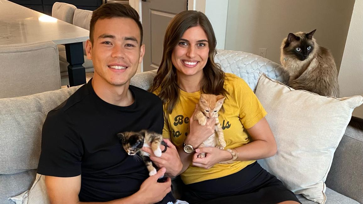 Sam Junqua and Emma Besier sitting on a couch holding foster kitten with a cat beside them on the armrest