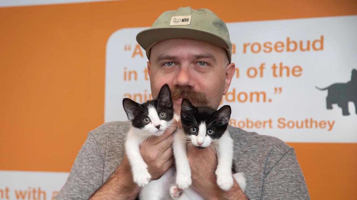 Foster Tyler Lisonbee wearing a hat and gray T-shirt, holding two black and white kittens in front of him