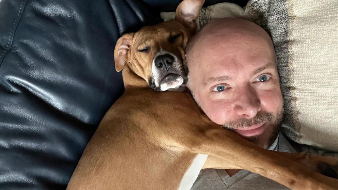 Volunteer David Sprague lying in a bed with a brown and white dog sleeping and snuggled next to him
