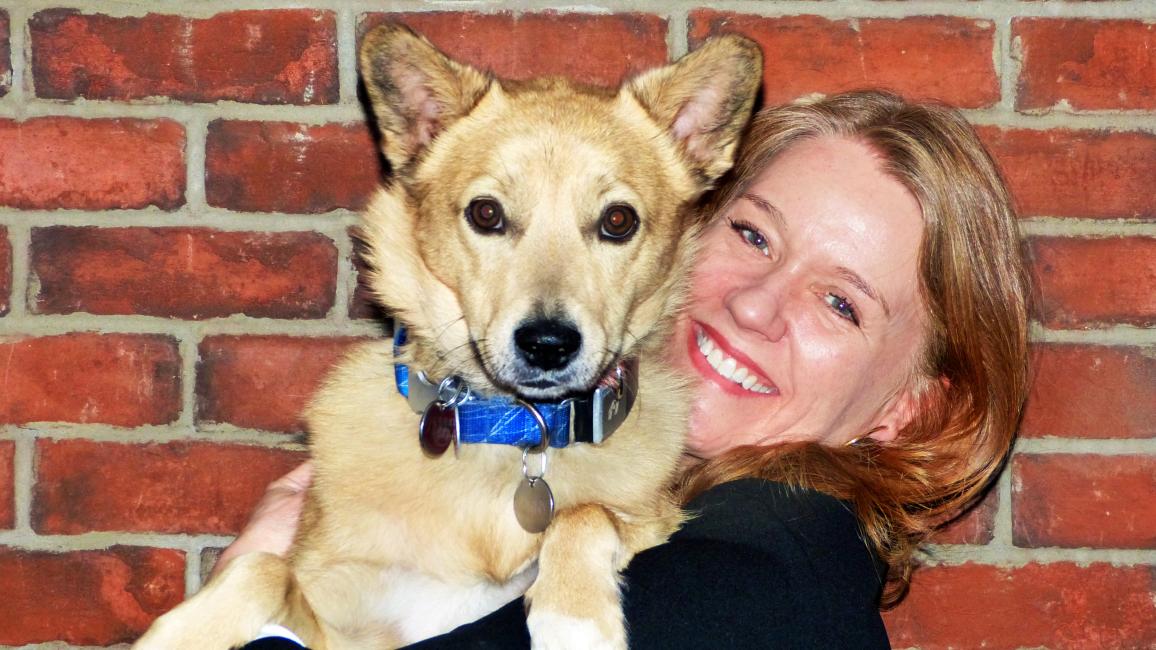 Smiling volunteer Vicki Williams holding Sunny her dog in front of a brick wall background