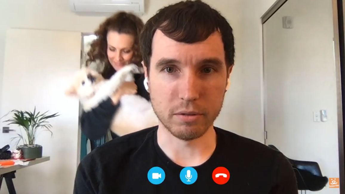 Screen shot of video of man, while a woman dances behind him cradling a dog