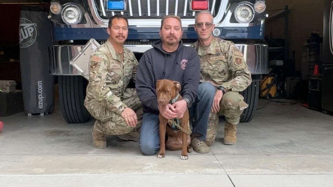 Ruthie the dog with three people (two in military uniforms) in front of a large vehicle