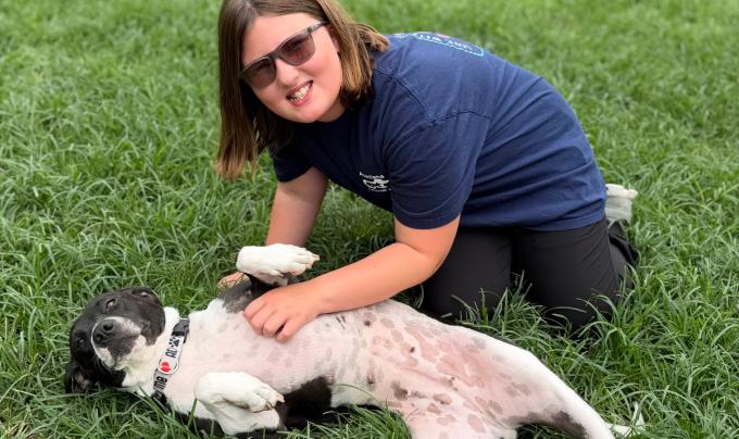 Person kneeling on the grass next to a dog who is lying on her back