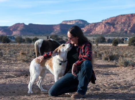 Emery holding Greenie the dog affectionately at the ranch with horses and rock formations in the background