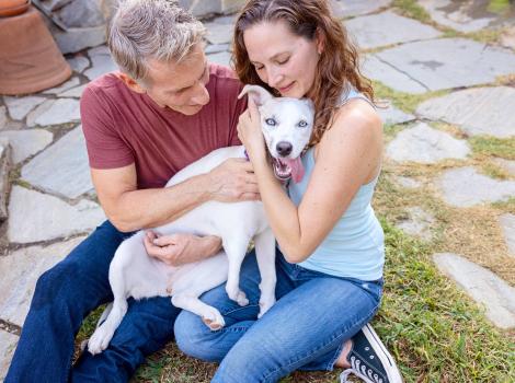 A couple hugging and petting a small white dog whose tongue is out