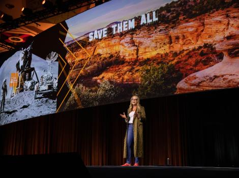 Best Friends CEO Julie Castle addressing the audience at the Best Friends National Conference with a large slide behind her featuring Angel Canyon with the words Save Them All