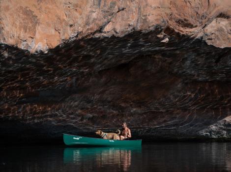 A person and dog in a green canoe on the water in front of a rock overcropping