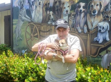Gary Slusher holding the Chihuahua-corgi mix he adopted in front of a mural featuring dogs in a covered wagon