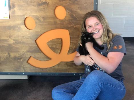 Woman holding a black cat sitting on the ground next to a Best Friends logo on the wall